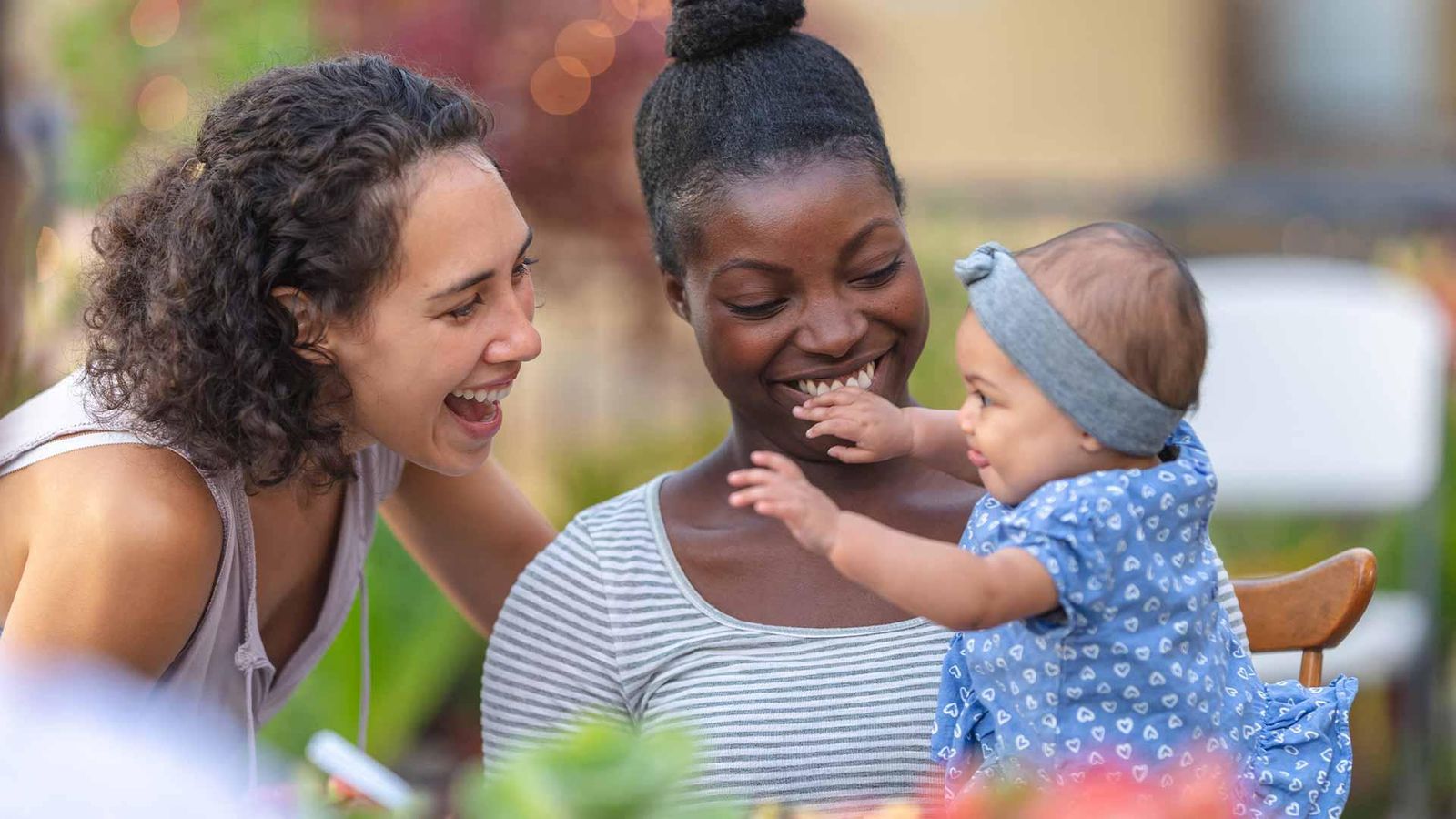 A young African American mom holds her adorable young daughter in her lap at an outdoor dining table.