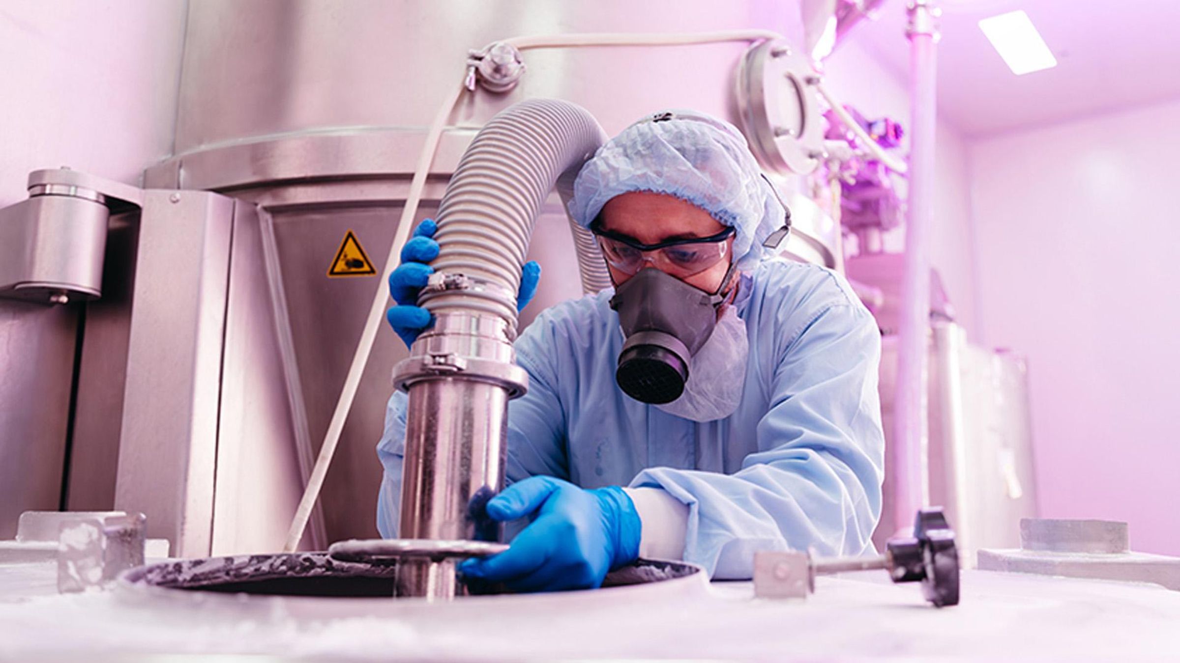 Technicien de laboratoire portant blouse bleue, gants, masque et lunettes de sécurité travaillant sur un appareil de filtration à spirale dans une salle de production pharmaceutique équipée de cuves et tuyauteries