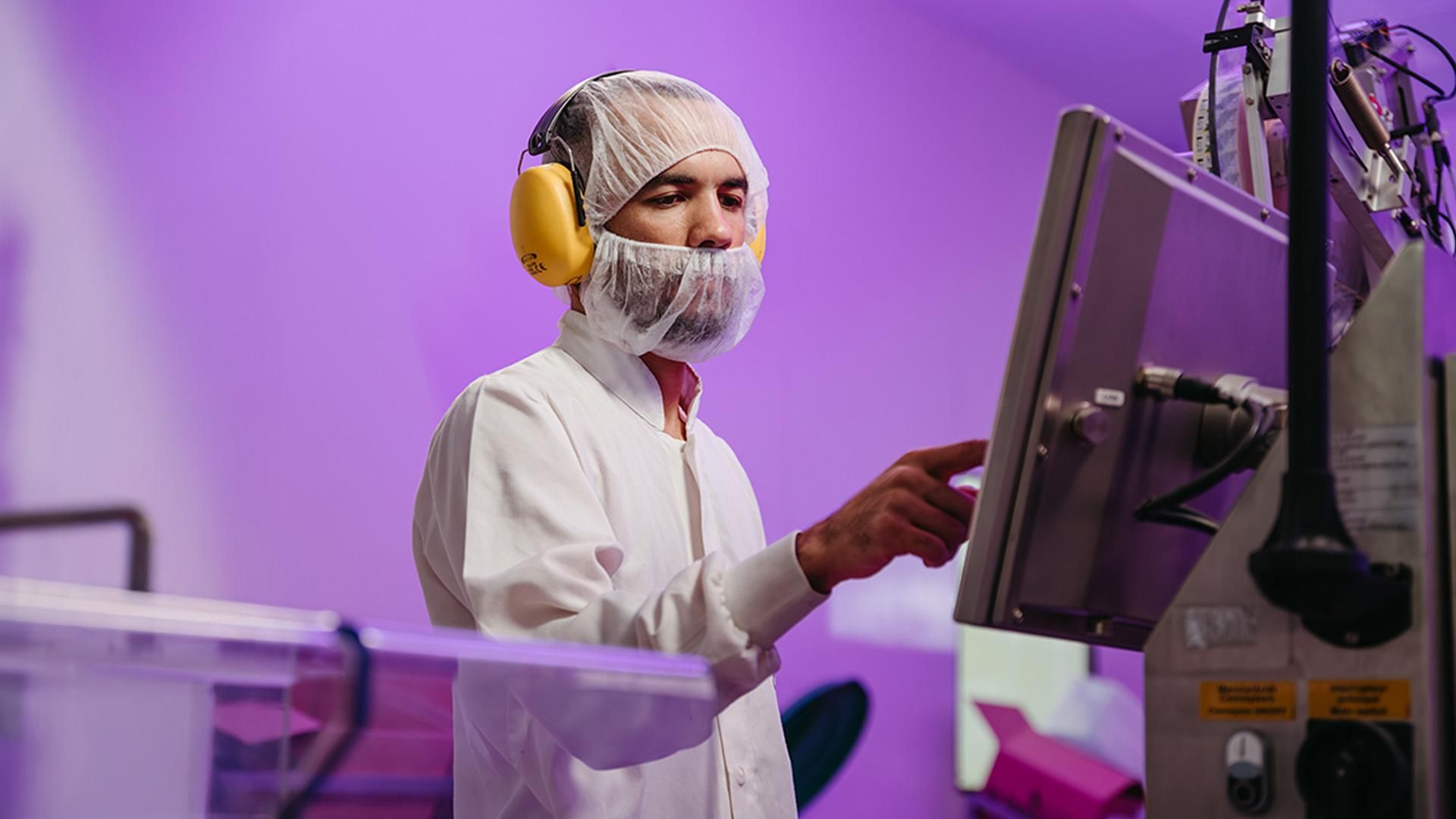 Opérateur en tenue de protection complète - blouse blanche, charlotte, masque et protection auditive jaune - pointant vers l'écran de contrôle d'une machine de production pharmaceutique dans un environnement stérile éclairé en violet
