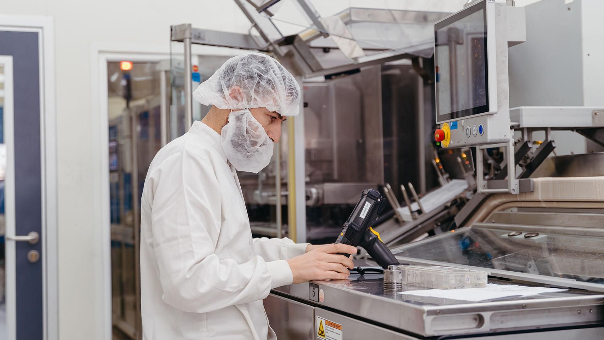 Technicien en blouse blanche, charlotte et masque facial manipulant un scanner à code-barres à poignée jaune devant un plan de travail en inox marqué du numéro 5 avec signalétique de sécurité, dans un environnement de production industrielle stérile