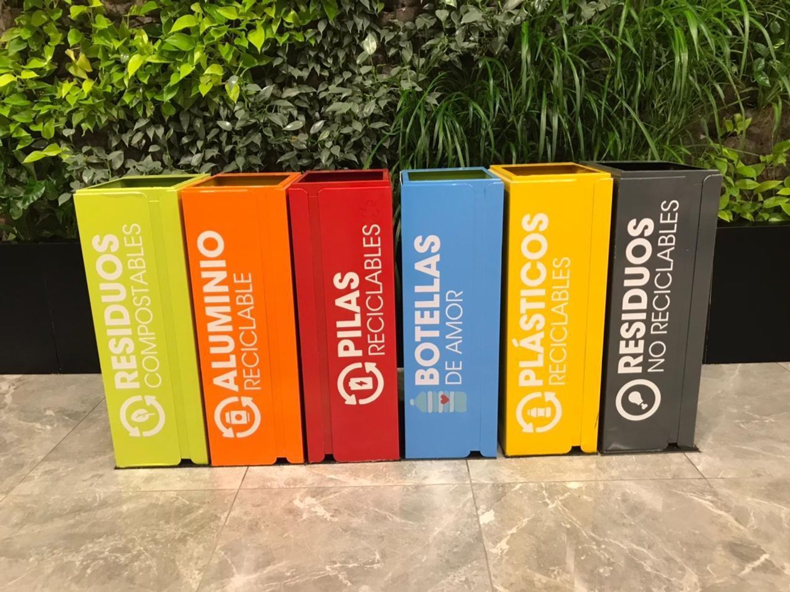 Seis cestos para desechos de colores verde, naranja, rojo, azul, amarillo y negro colocados dentro de una oficina, frente a una pared con plantas verdes.