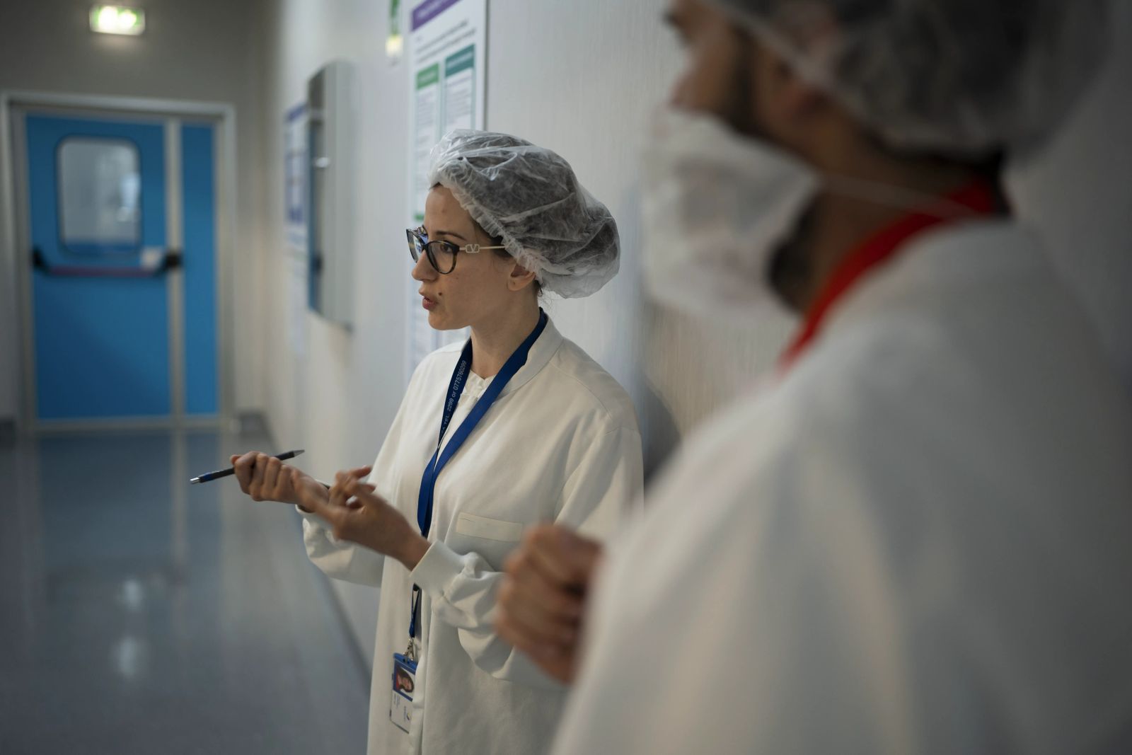 Eine Frau im Vorder und ein Mann im Hintergrund, in weißen Laborkitteln und Haarnetzen, unterhalten sich im Flur eines Labors