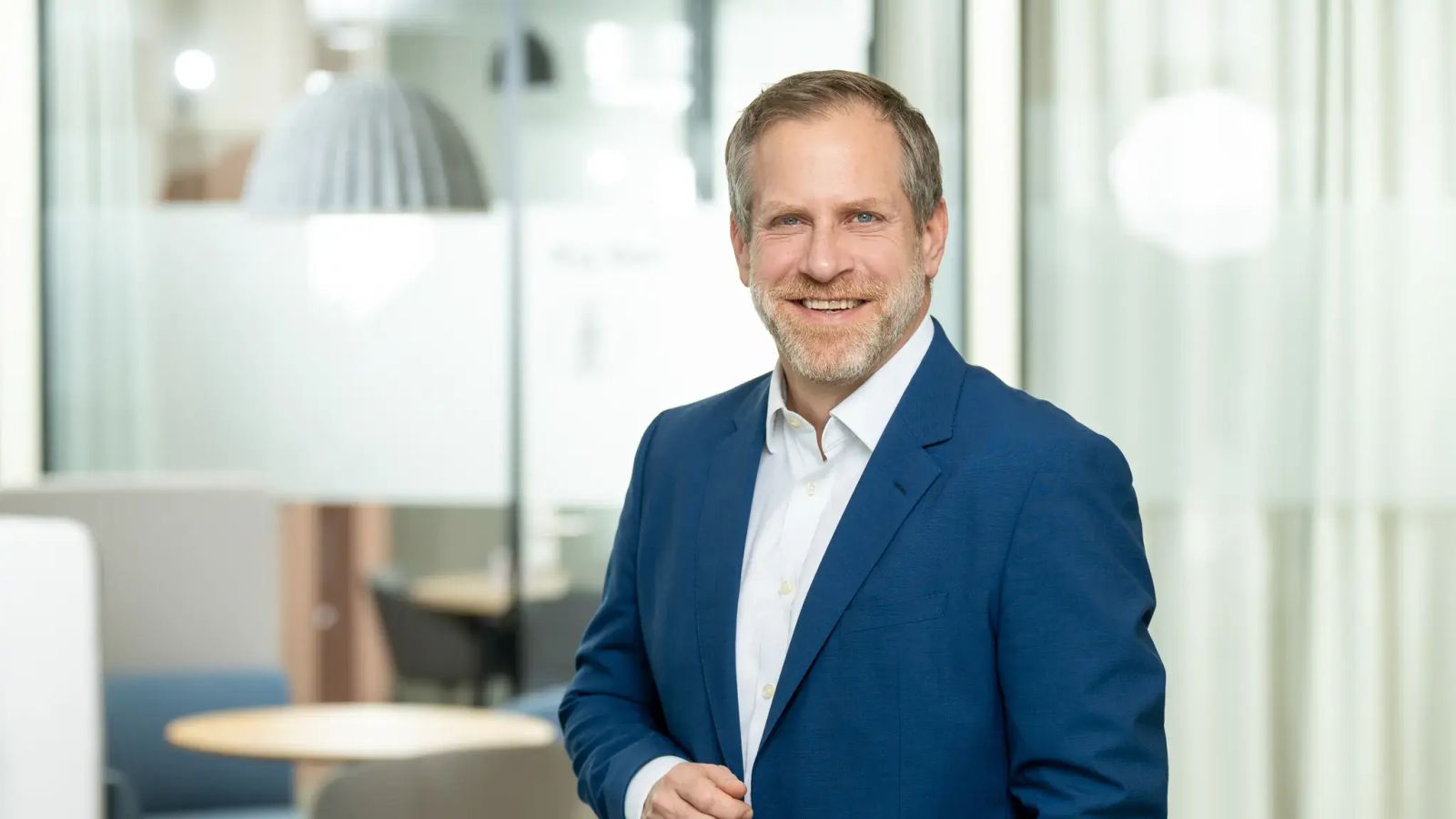 Mann im weißen Hemd und blauen Sakko, lächelt professionell in die Kamera und vermittelt Kompetenz.