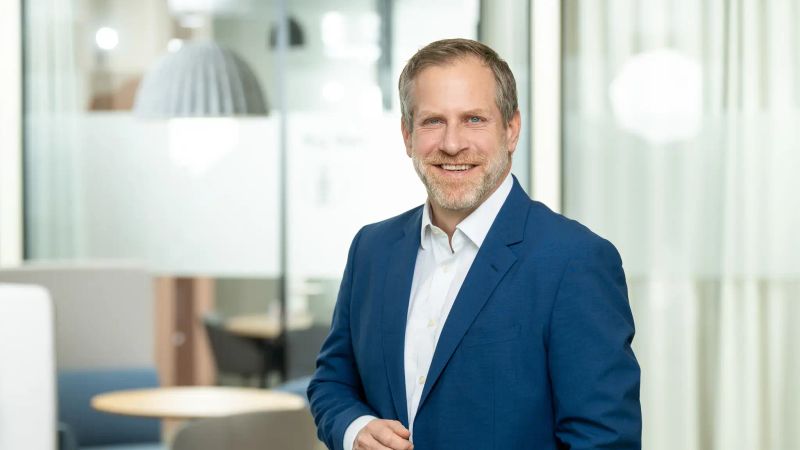 Mann im weißen Hemd und blauen Sakko, lächelt professionell in die Kamera und vermittelt Kompetenz.