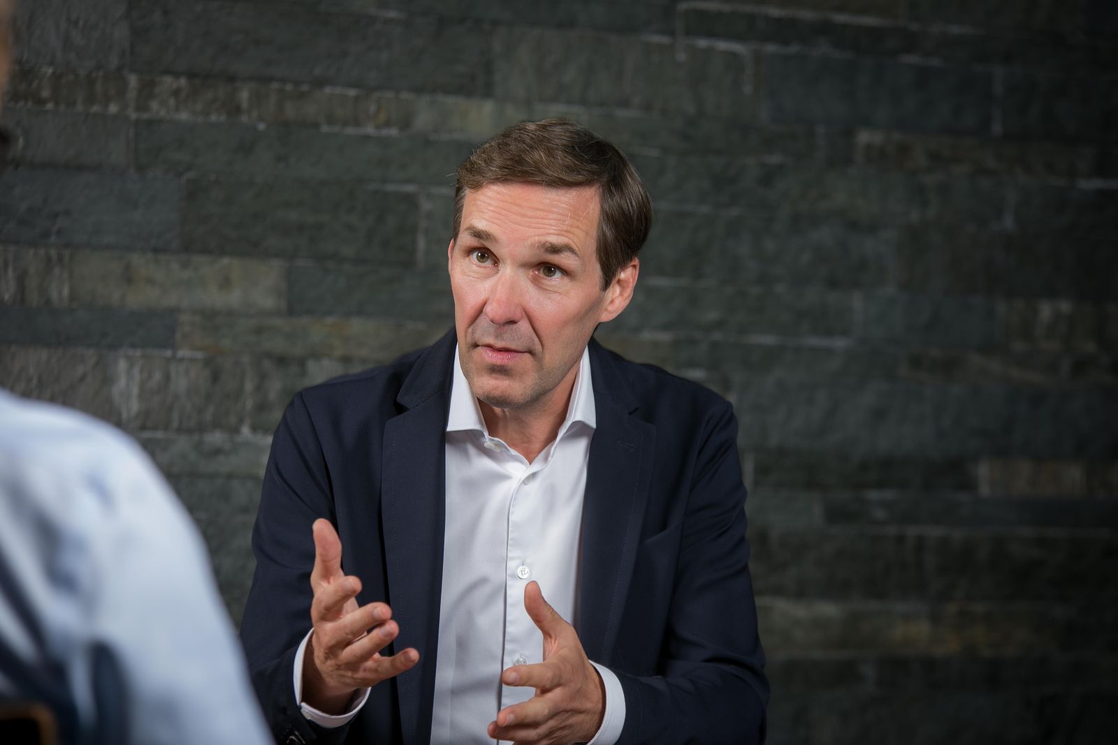Ein Mann spricht gestikulierend in einem dunklen Anzug vor einer grauen Steinwand.
