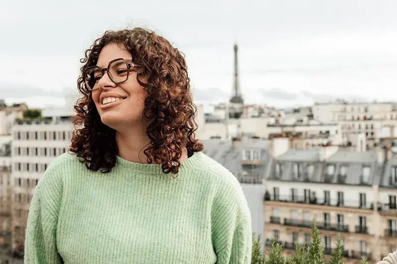 Eine Frau mit schulterlangen lockigen Haaren, Brille und Pullover lächelt zur Seite, im Hintergrund sind Häuser und der Eiffelturm zu sehen | Caption: fc Zoe Savitz