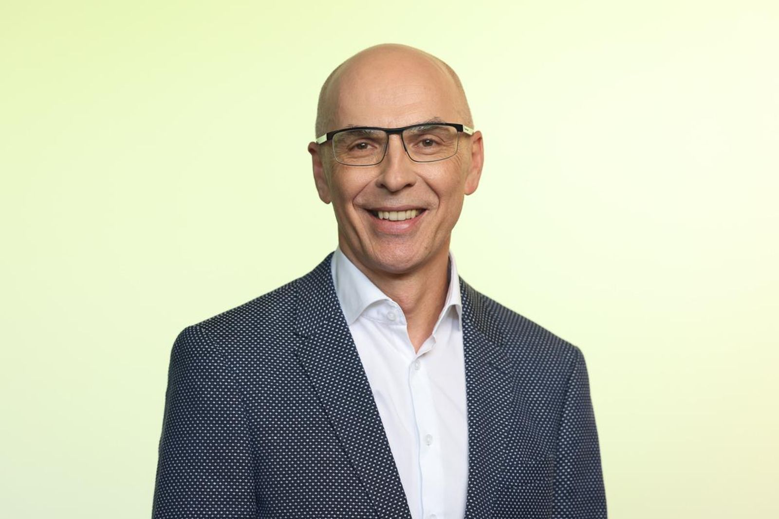 Ein Mann lächelt, trägt eine Brille, Glatze und ein dunkelblaues, gepunktetes Sakko auf einem gelben Hintergrund.