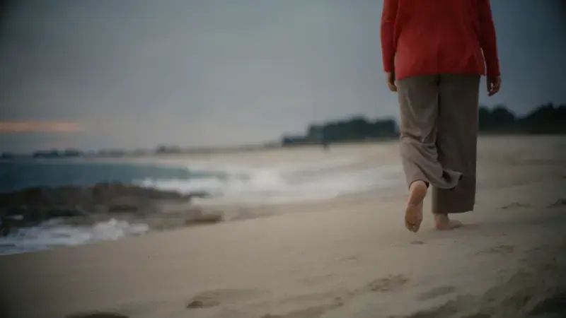 Eine Frau hinterlässt Fußspuren im Sand, als sie mit barfuß entlang des bewölkten Strandes von der Kamera weggeht | Caption: fc Satellite My Love