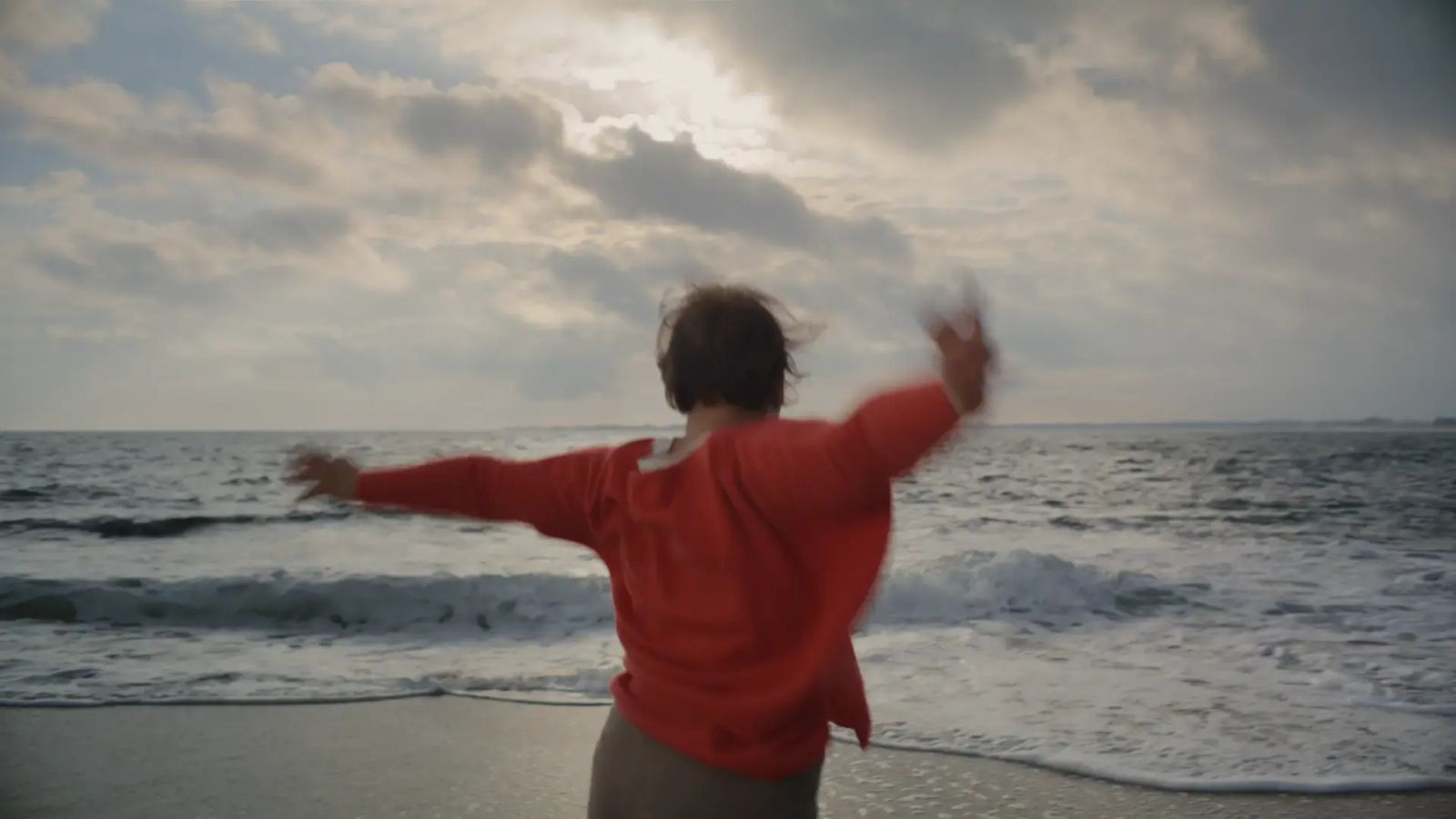 Frau, von hinten am Strand tanzend, mit erhobenen Armen und rotem Pullover. Im Hintergrund sind Meereswellen und Sand.