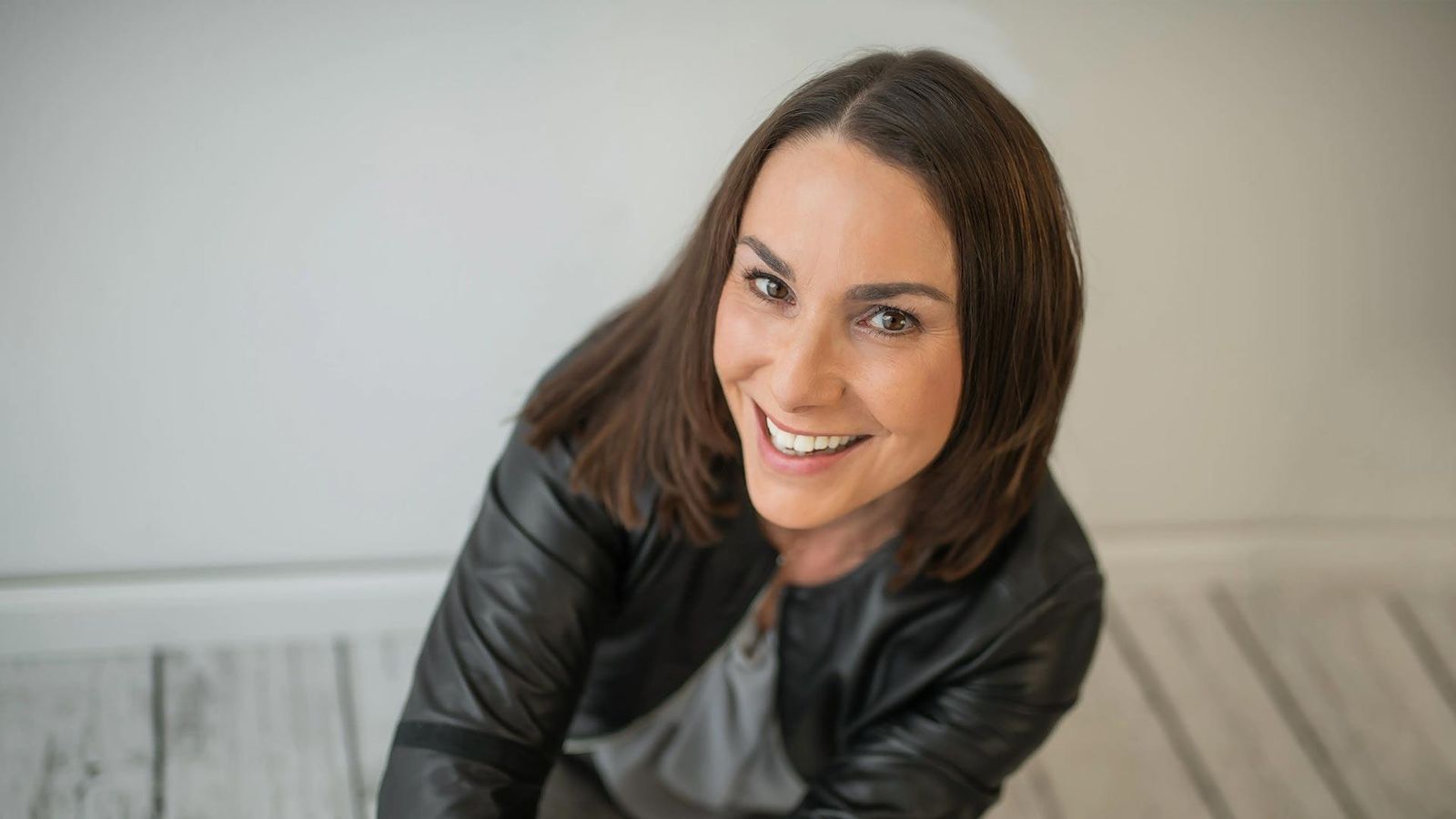Eine Frau lächelt strahlend in die Kamera, trägt eine schwarze Lederjacke und hat schulterlanges, glattes, braunes Haar.