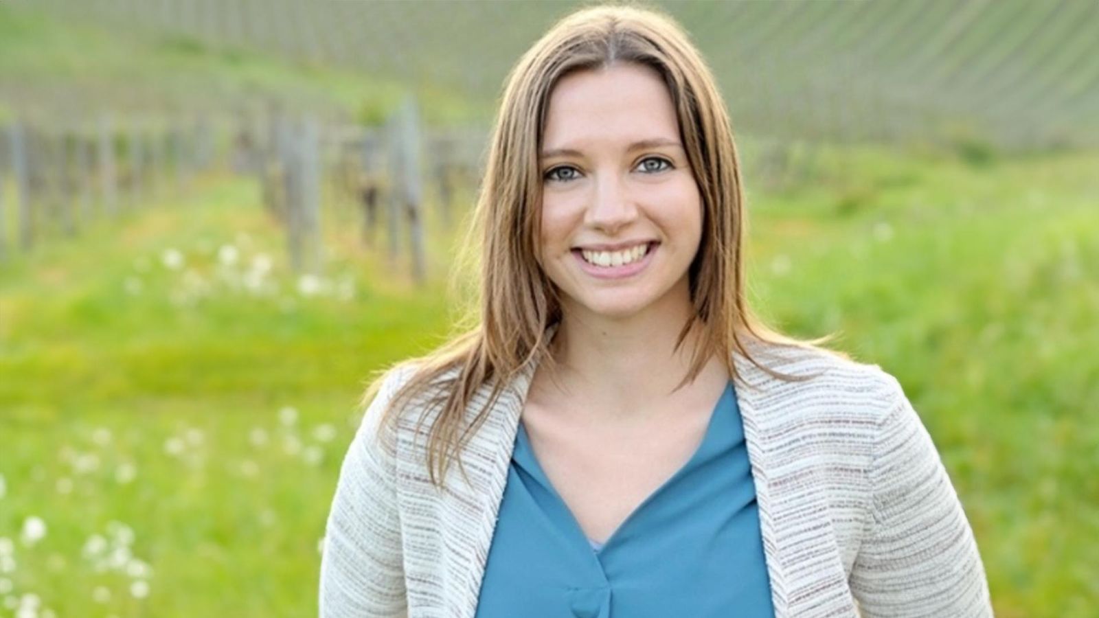 Eine junge Frau lächelt in einer grünen Landschaft vor einem Weinberg, mit schulterlangem, hellbraunem Haar und blauer Bluse.
