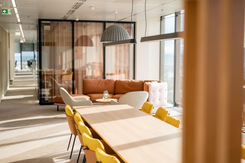Helles Büro mit großen Fenstern, gelben Stühlen, oranger Couch und gemütlicher Sitzecke. Eine warme, offene Atmosphäre.