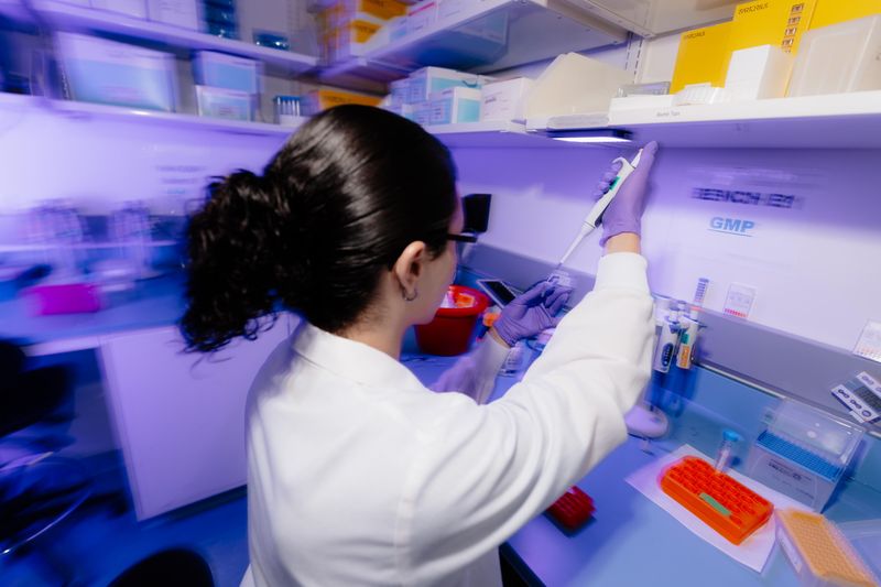 Scientifique avec des cheveux noirs bouclé, travaille avec des échantillons avec des gants violets, une tenue de laboratoire et des lunettes de sécurité.