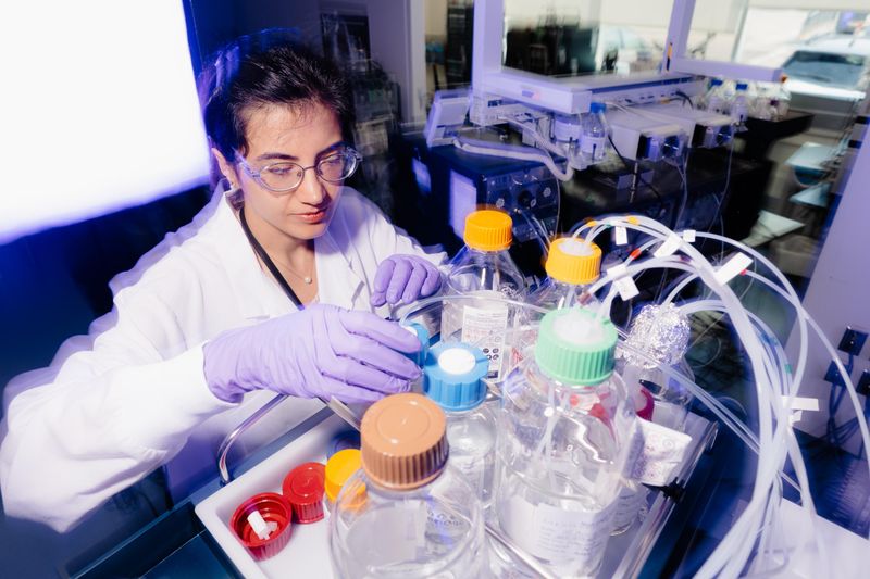 La scientifique a de longs cheveux noirs bouclés, porte des lunettes de sécurité et une blouse de laboratoire blanche, et tient du verre dans une pièce éclairée en violet