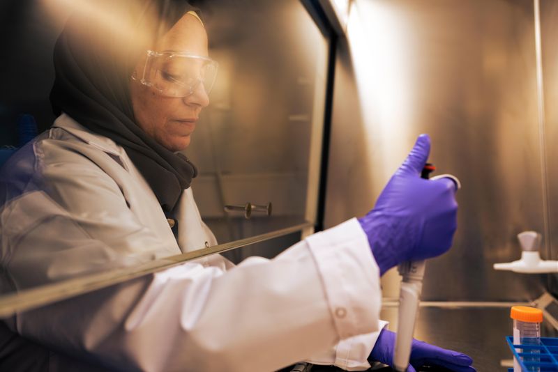 Nadin Al Shukor prépare des échantillons sous une hotte, portant des lunettes de sécurité, des gants violets, une blouse blanche et un foulard