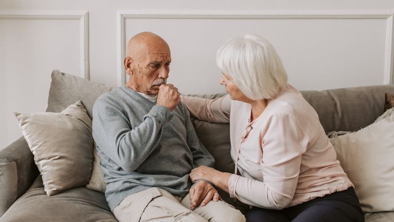 Adultos mayores sentados en un siñón, uno tosiendo y la otra persona atenta a él.