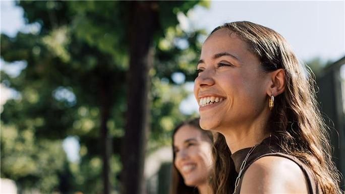 Two women laughing together outdoors in a sunny park with green trees in the background