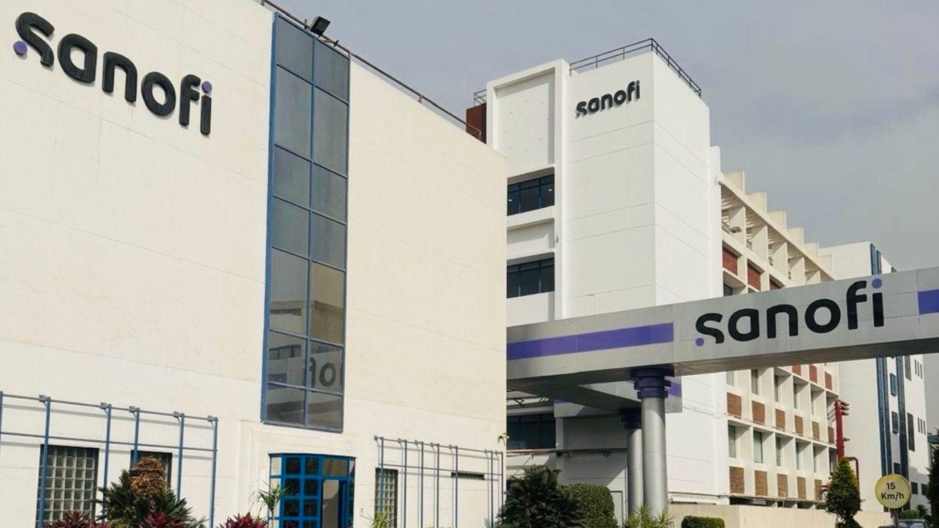 Exterior view of a contemporary multi-story Sanofi office building with white and blue façade, featuring the Sanofi logo on both the building front and entrance canopy, surrounded by manicured greenery and a 15 km/h speed limit sign.