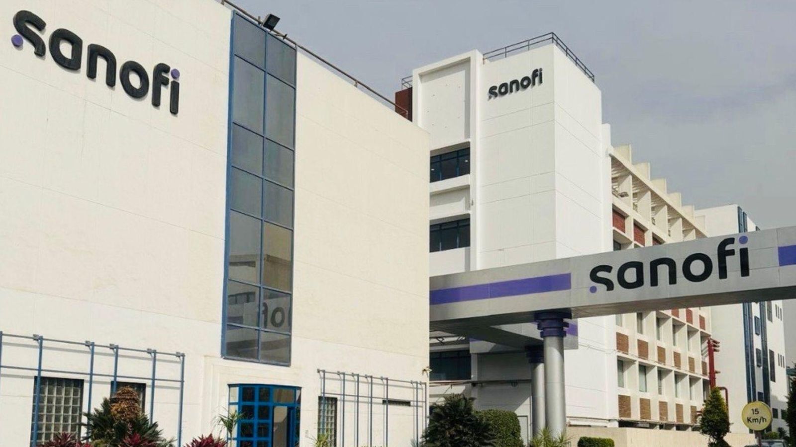 Exterior view of a contemporary multi-story Sanofi office building with white and blue façade, featuring the Sanofi logo on both the building front and entrance canopy, surrounded by manicured greenery and a 15 km/h speed limit sign.