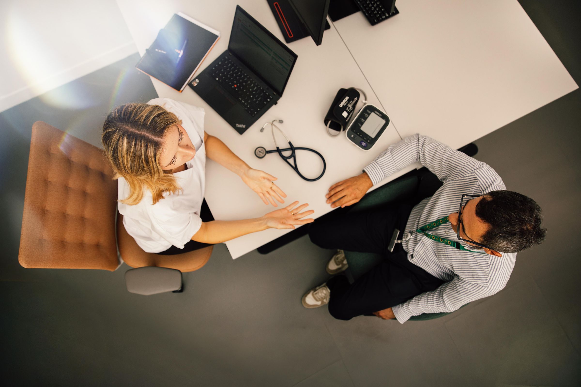Overhead view of a desk with a healthcare professional and a patient in conversation, stethoscope and laptop visible.