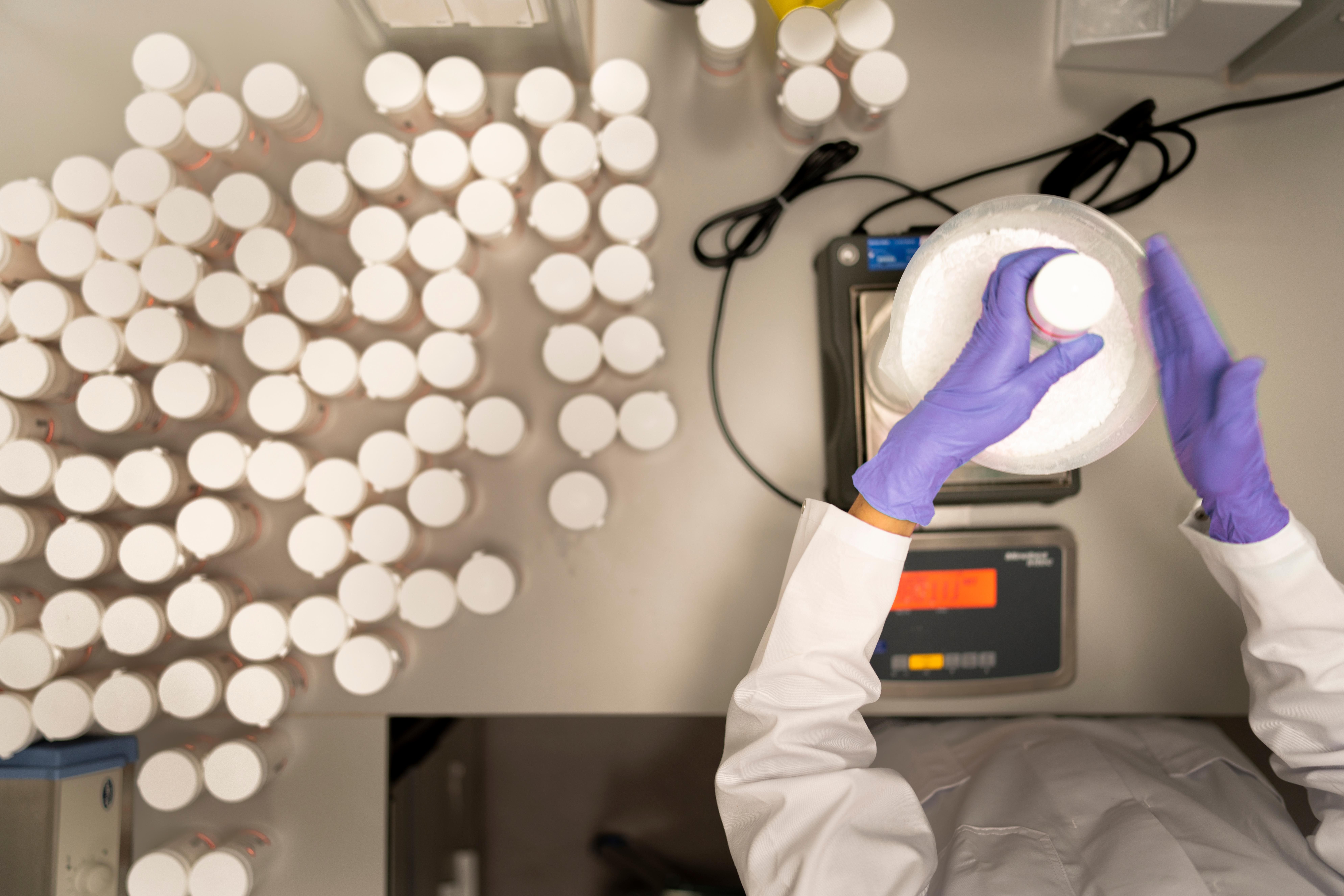 An overhead view of a technician weighing a large plastic container of white powder, with hundreds of smaller containers nearby