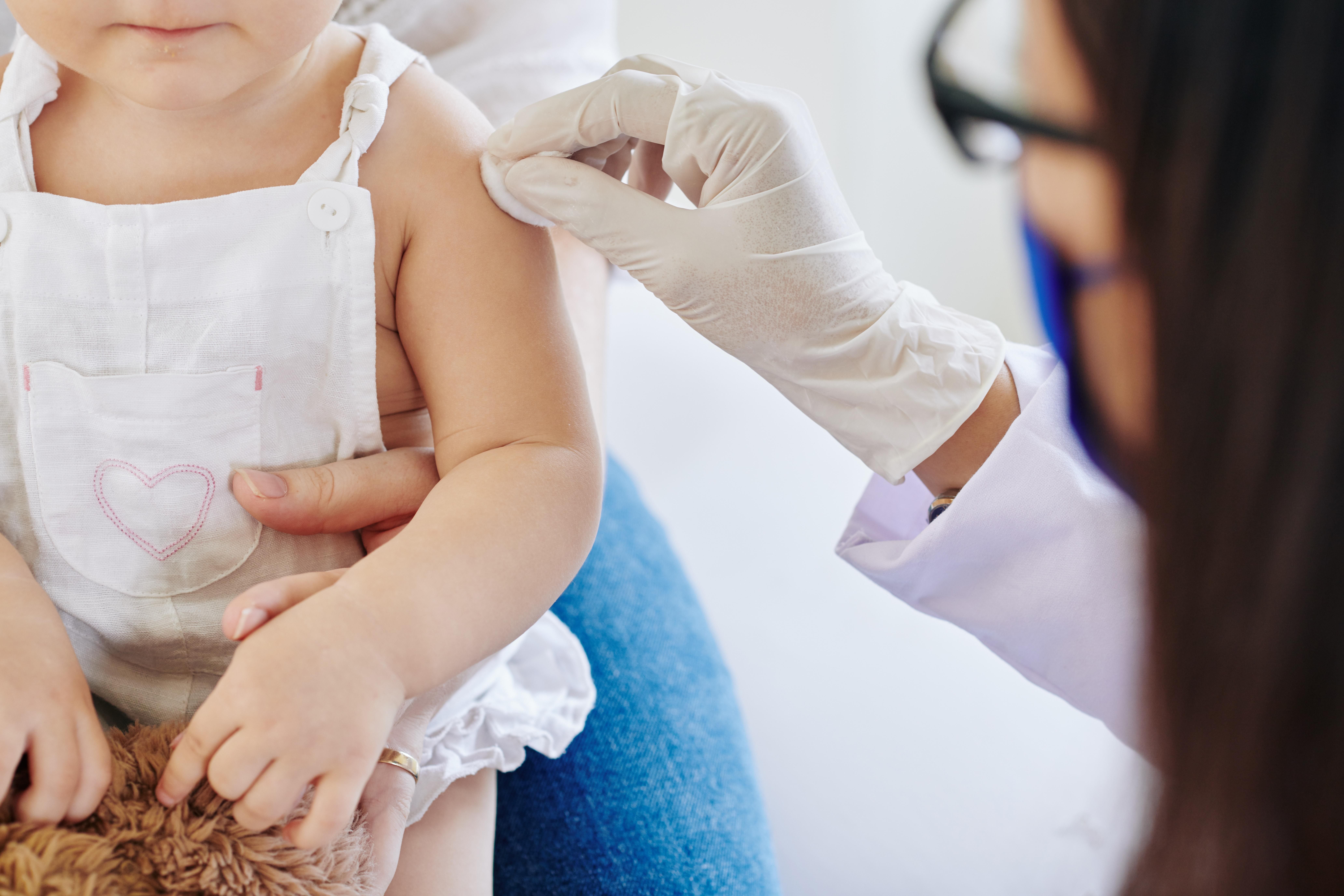 Profesional médico con bata blanca, guantes y mascarilla administrando una vacuna en el brazo de un niño que viste un vestido blanco con corazón rosa y sostiene un peluche marrón, en un entorno clínico
