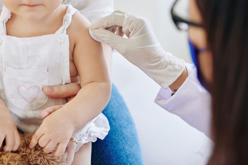 Profesional médico con bata blanca, guantes y mascarilla administrando una vacuna en el brazo de un niño que viste un vestido blanco con corazón rosa y sostiene un peluche marrón, en un entorno clínico