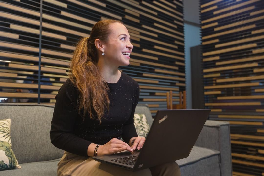 Mujer sonriente sentada en un sofá con una computadora portátil en el regazo, mirando hacia alguien fuera del encuadre