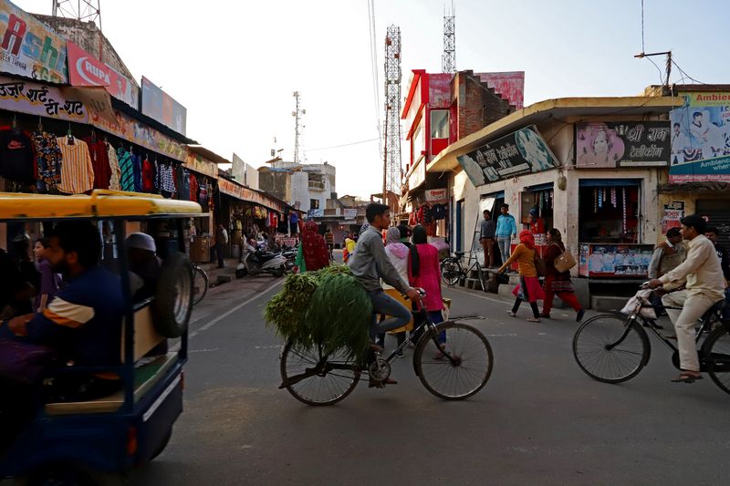 Оживлённая сцена городского рынка: по улице едут велорикша на жёлто-синем трёхколёсном велосипеде с пассажиром и велосипедист с охапкой зелени. По обеим сторонам улицы расположены магазины и лавки с одеждой и навесами. Многоэтажные здания из бетона и кирпича, над улицей протянуты электрические провода. Люди в традиционной и современной одежде заняты повседневными делами.