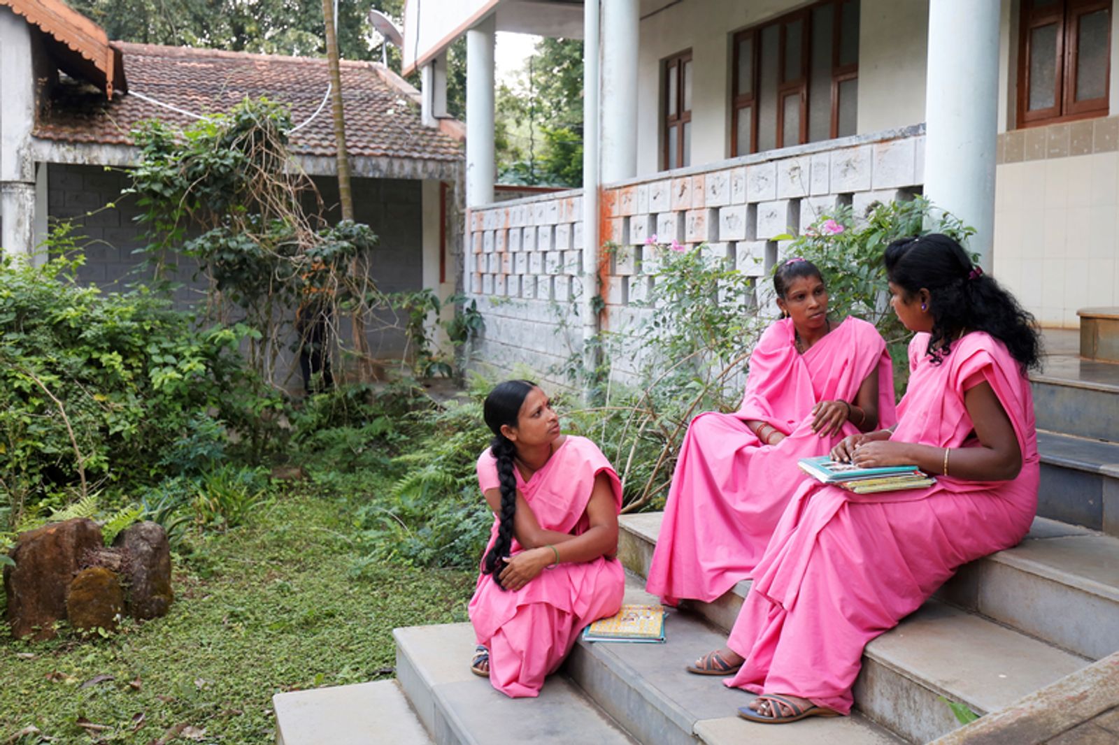 Три женщины в розовых сари беседуют на ступенях здания в саду, одна из них держит стопку книг