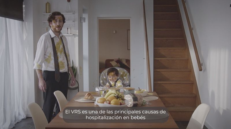 Imagen de una cocina donde el padre lleva puesta una a camisa blanca manchada por la comida de su hijo mientras este último, sentado en una silla con la comida delante, mira sonriente a la cámara.
