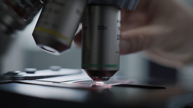 Plano cerrado de un microscopio en un laboratorio donde se visualiza una mano de fondo manipulándolo.