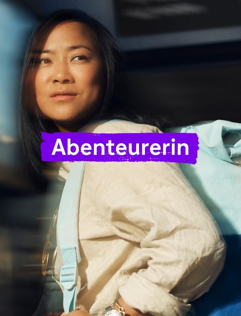 Eine Frau mit gelbem Shirt und blauem Rucksack blickt in die Ferne mit einem verträumten Ausdruck. Überlagerter Text lautet: „Abenteurerin.“​