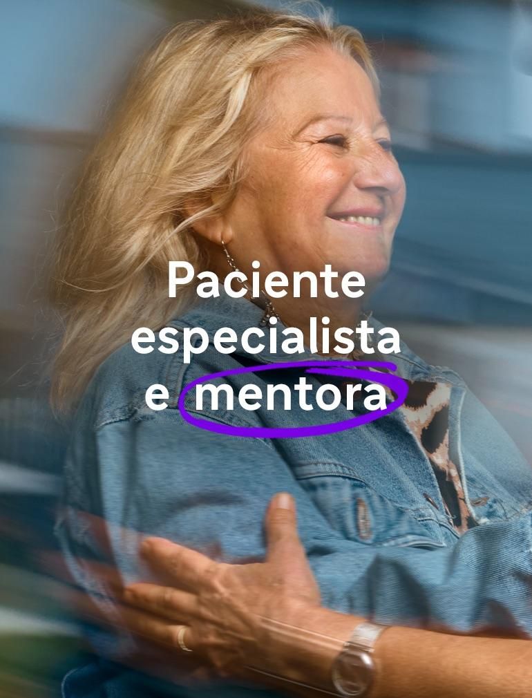 Uma mulher sorridente de jaqueta jeans azul estende os braços para abraçar outra mulher usando óculos. Um texto sobreposto diz: ‘Paciente-especialista e mentora’.