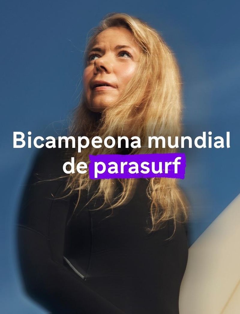 Una mujer con un traje de neopreno negro sostiene una tabla de surf bajo un cielo azul y soleado, mirando hacia el horizonte.