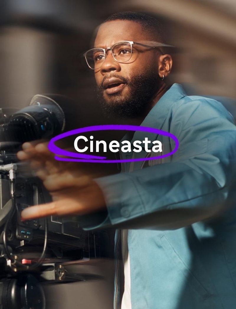 Un hombre con gafas y una chaqueta azul opera con una cámara de vídeo y se muestra concentrado mientras graba; extiende una mano hacia delante haciendo un gesto.