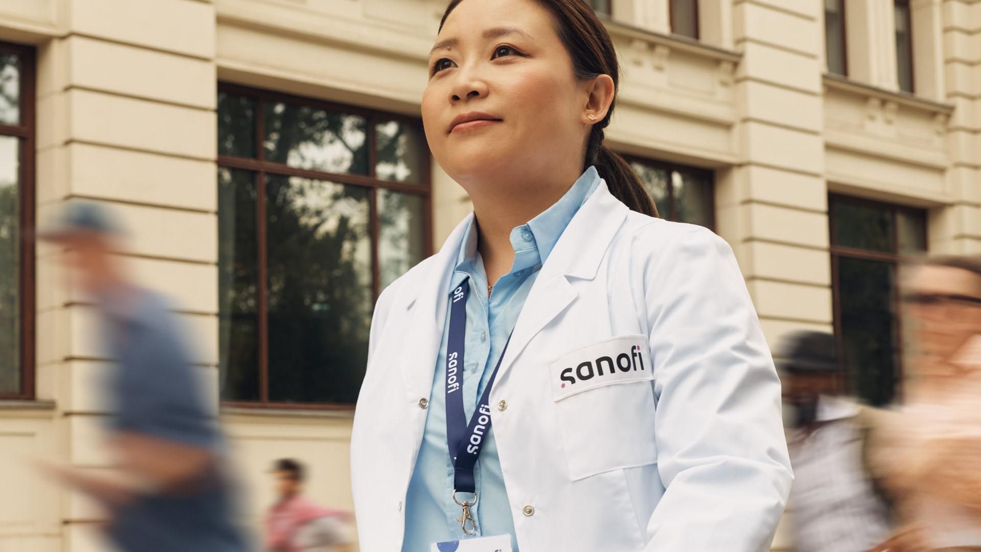 A woman in a white Sanofi lab coat and company badge outside of a building, moving forward while confidently and positively looking ahead. Several people are running alongside her in the background.​