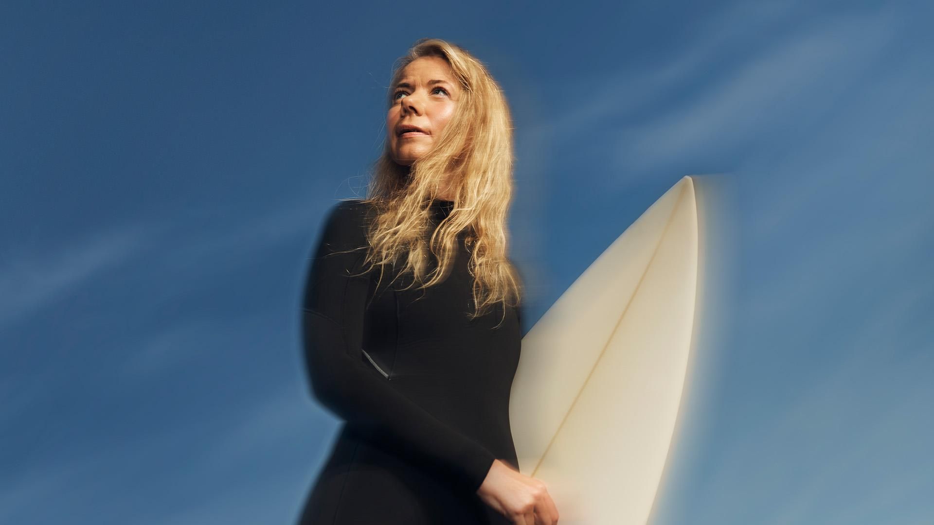 A woman wearing a black wetsuit holds a surfboard under blue skies and sunshine while looking out to the horizon with a hopeful and determined expression.​