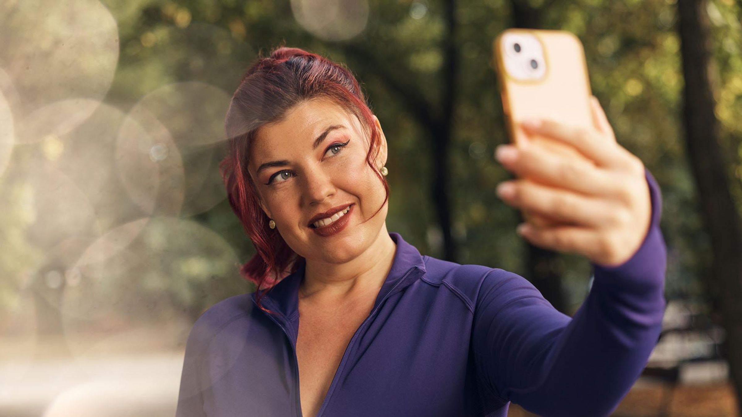 A smiling woman wearing a purple athletic top, holds up a smartphone to take a selfie outdoors. The background is blurred green foliage, and the sun is shining.