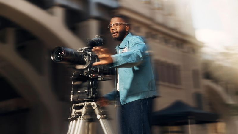 A man wearing glasses and a blue jacket operates a video camera and looks focused while filming, his hand is outstretched in front of him as he gestures. 