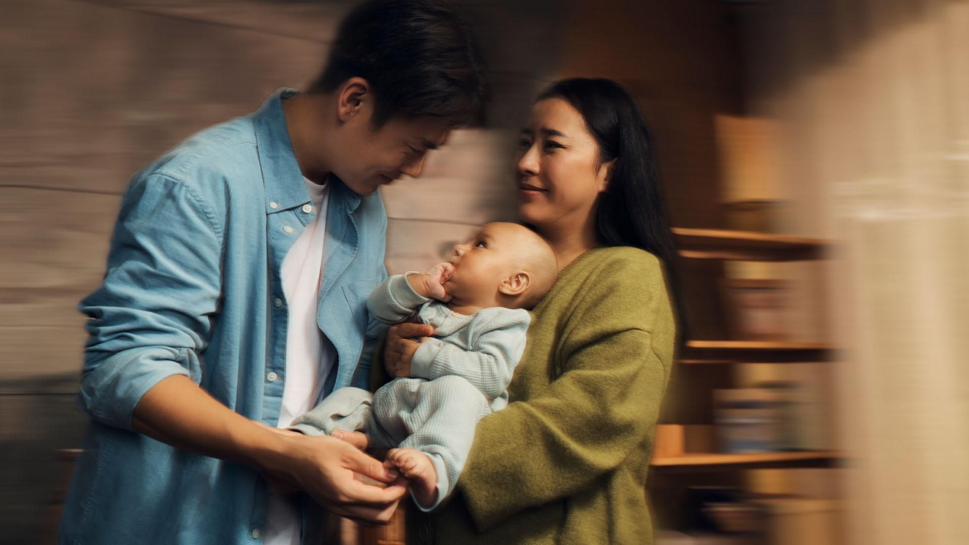 A man in a blue shirt and a woman in a green sweater hold a small baby together in a warmly lit room. The baby has its hand in its mouth, looking up at the man lovingly.​