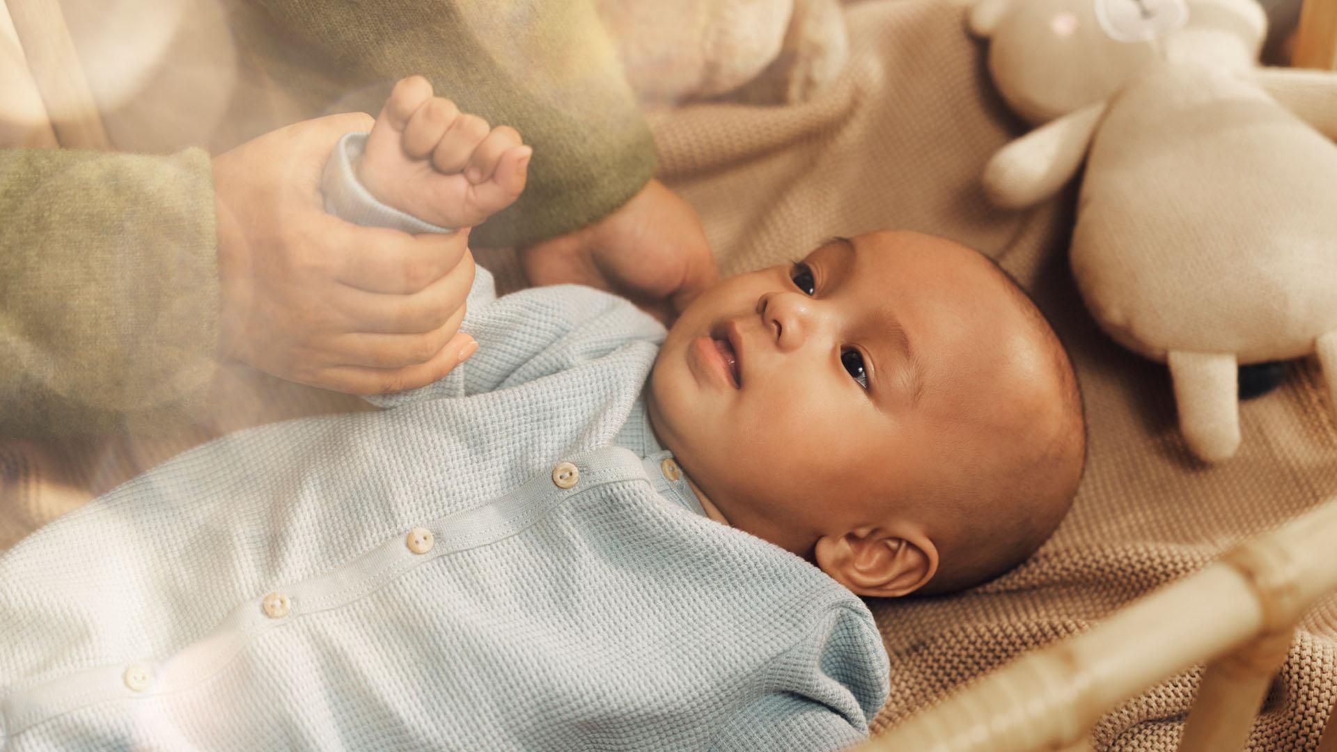 A small baby dressed in light blue clothing on a blanket in a crib is looking up. A woman’s hands are cradling the baby’s head and gently holding its arm, with a stuffed toy  nearby.​