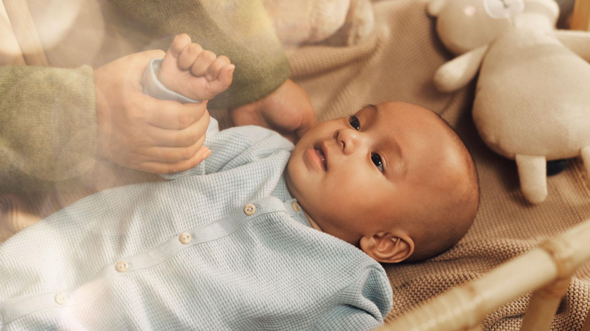 A small baby dressed in light blue clothing on a blanket in a crib is looking up. A woman’s hands are cradling the baby’s head and gently holding its arm, with a stuffed toy  nearby.