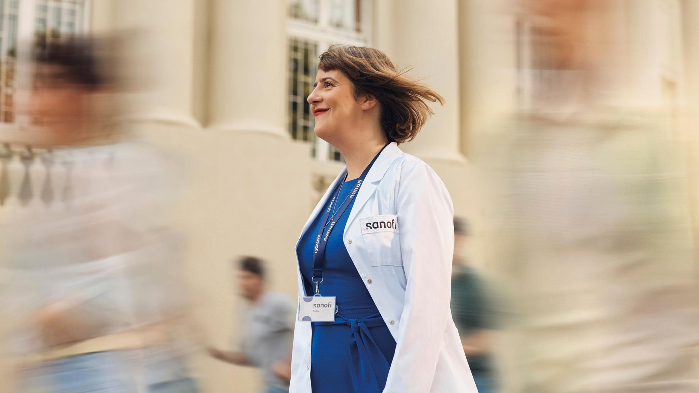 A smiling woman wearing a white Sanofi lab coat and company badge smiles while walking outdoors. Several people are running alongside her in the background, they are blurred in motion.