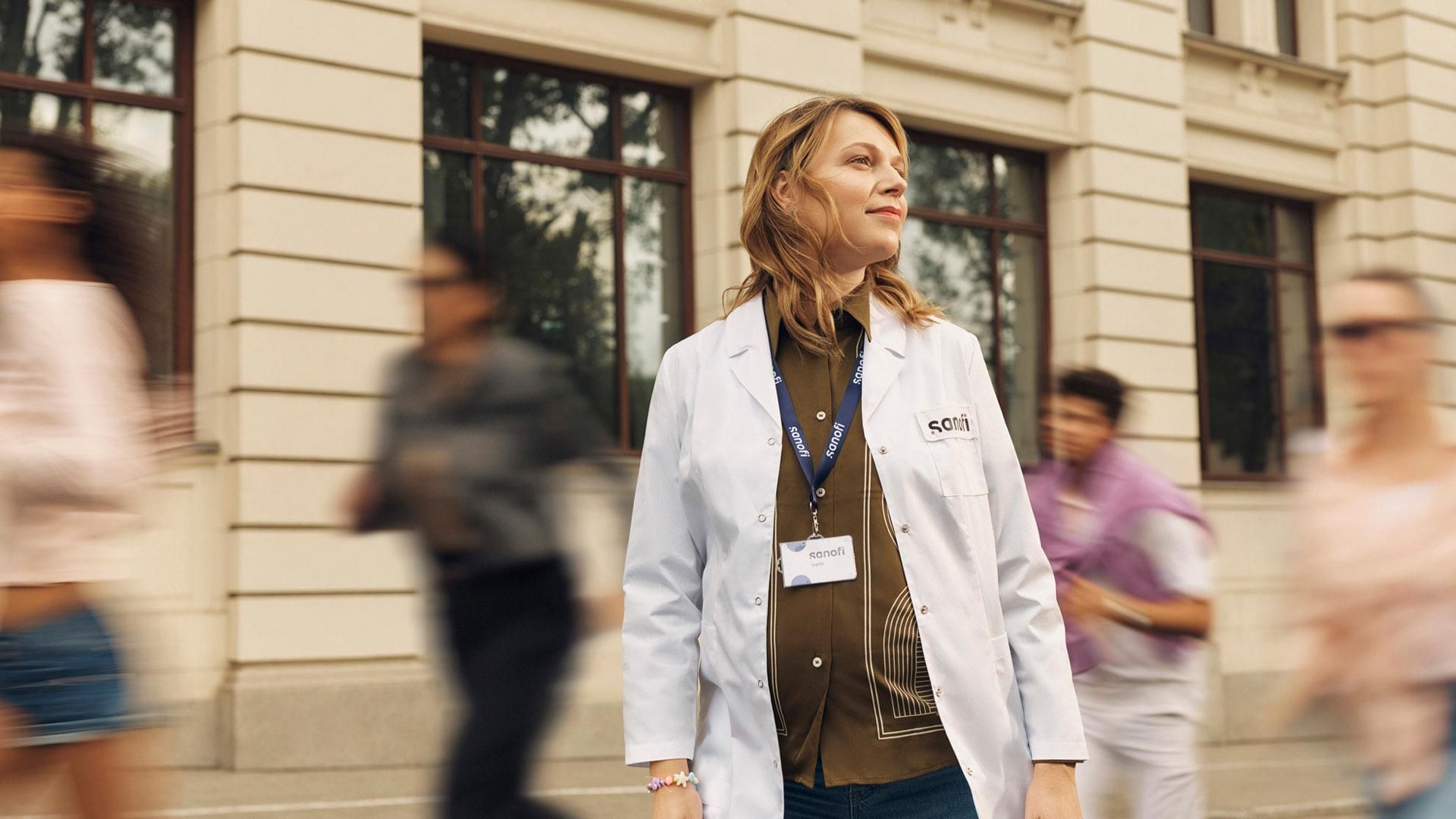 A woman in a white Sanofi lab coat and company badge looks ahead with an optimistic expression while standing outside a building. Several people are running alongside her in the background, they are blurred in motion.​