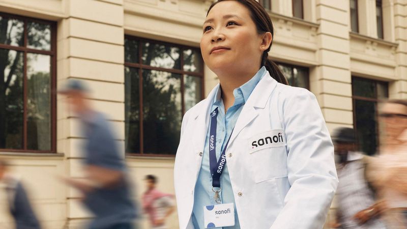 A woman in a white Sanofi lab coat and company badge outside of a building, moving forward while confidently and positively looking ahead. Several people are running alongside her in the background.