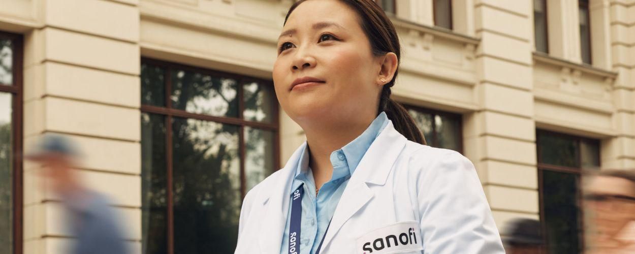 Une femme portant une blouse de laboratoire blanche Sanofi et un badge de l’entreprise, à l’extérieur d’un bâtiment, avance en regardant devant elle avec assurance et optimisme. Plusieurs personnes courent à ses côtés à l’arrière-plan.​