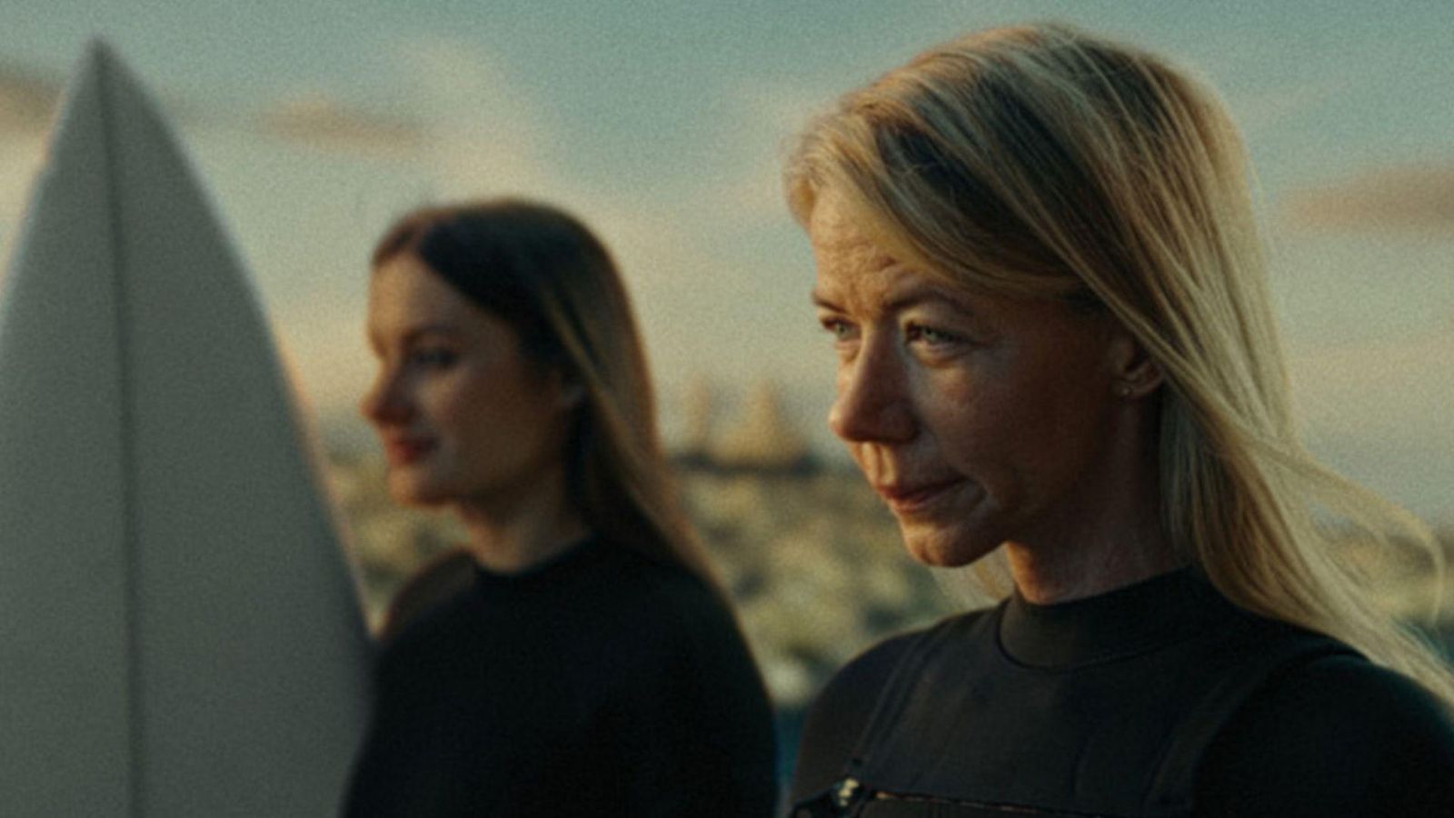 Two women in black wetsuits standing on a beach with a surfboard, ready for surfing