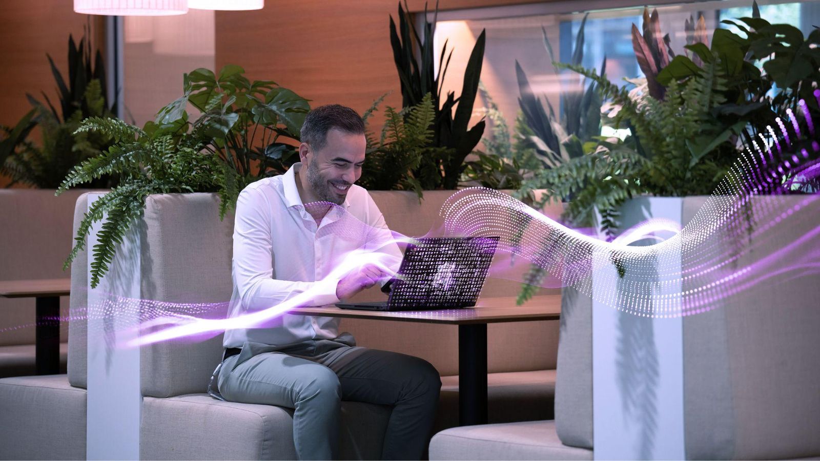 Une personne assise dans un espace de travail moderne rempli de plantes, travaillant sur un ordinateur portable à une table de type alcôve. Des lumières suspendues éclairent doucement la zone, et un graphique numérique ondulant de couleur violette traverse le premier plan.