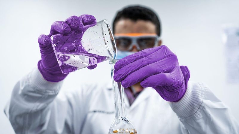 Un hombre con guantes morados, bata blanca y gafas de protección vierte líquido transparente de un vaso de precipitados a un matraz.